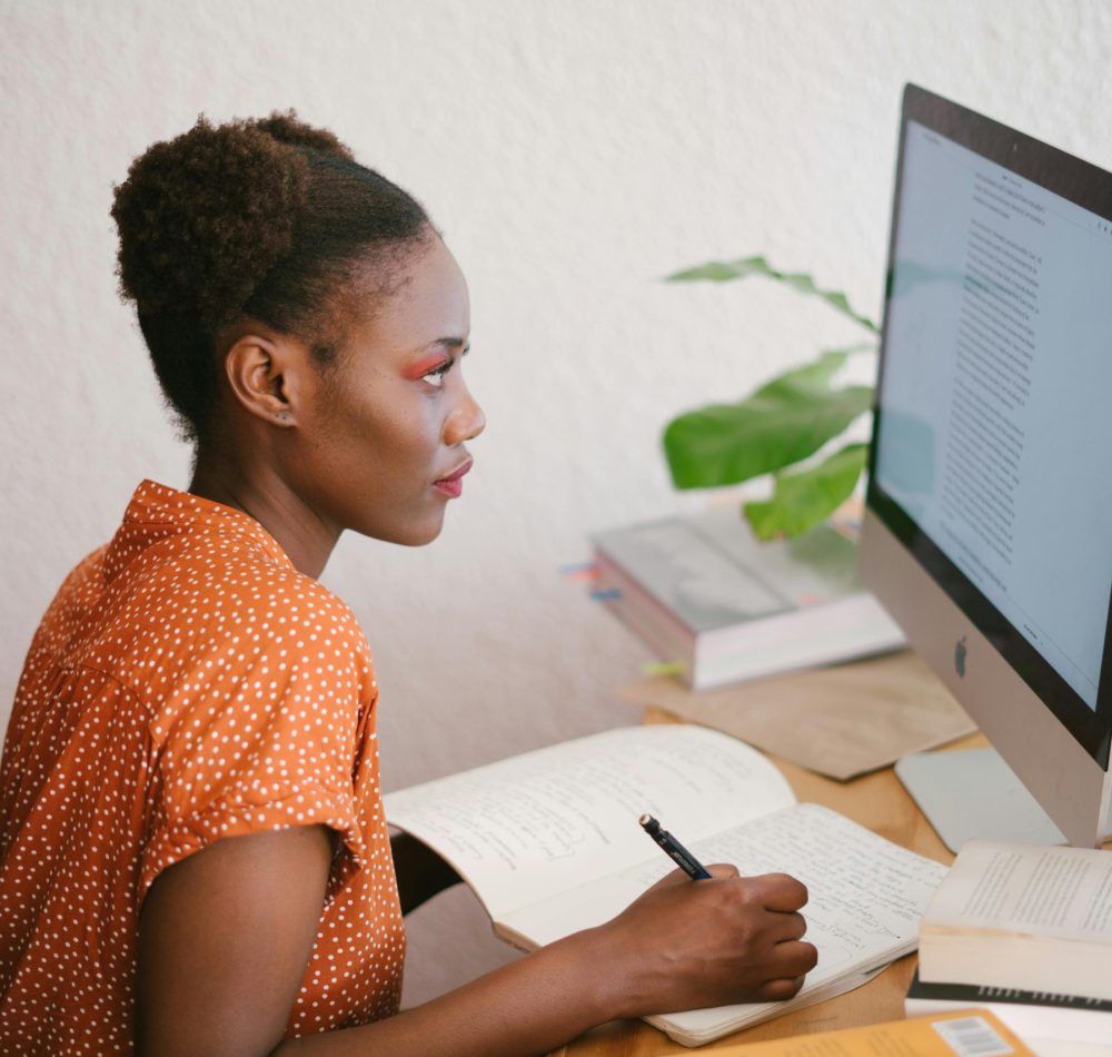 A focused young woman studying with books and a computer at her home office desk.
