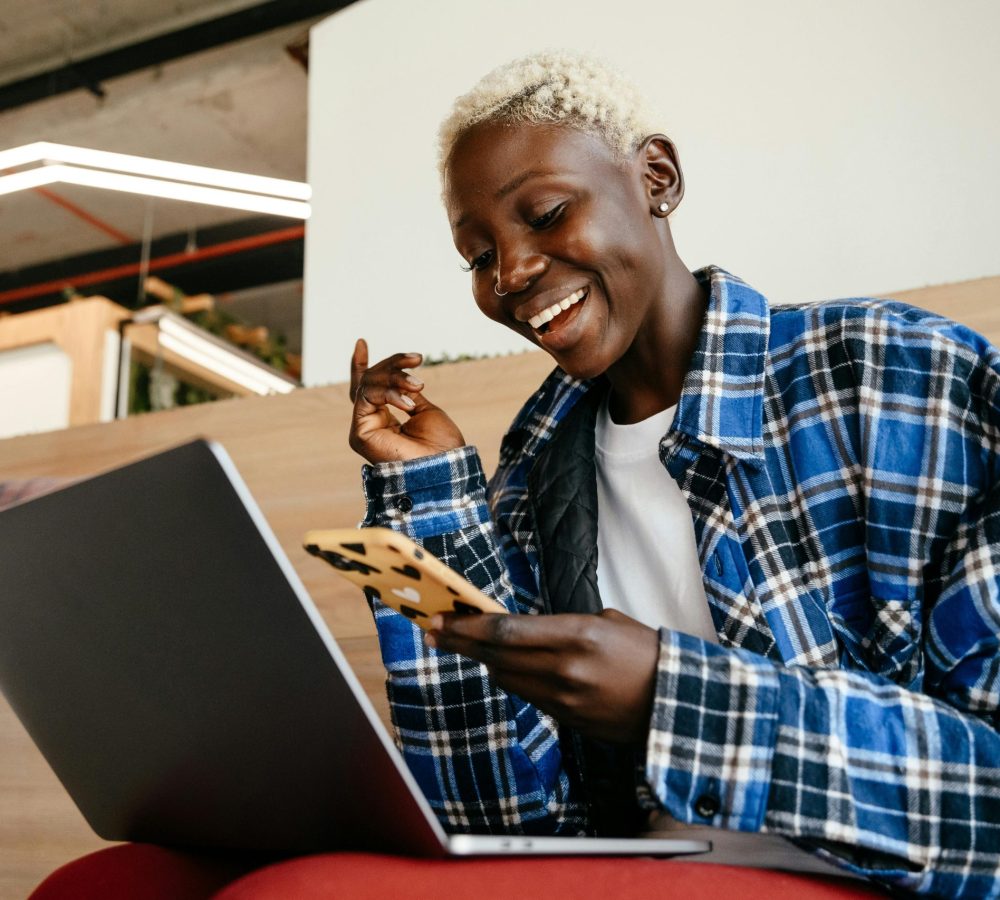 From below of cheerful African American female smiling and texting message on mobile phone while working with netbook