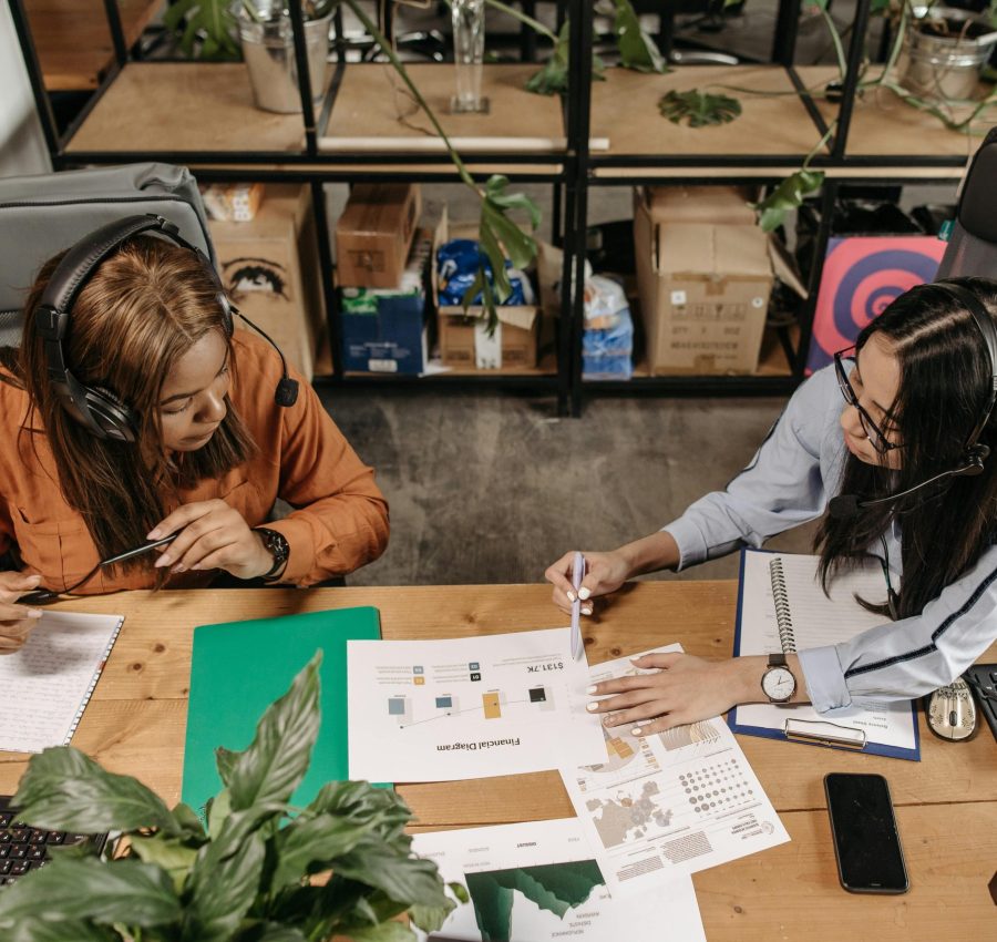 Two women wearing headsets discuss work at a shared desk in an office.